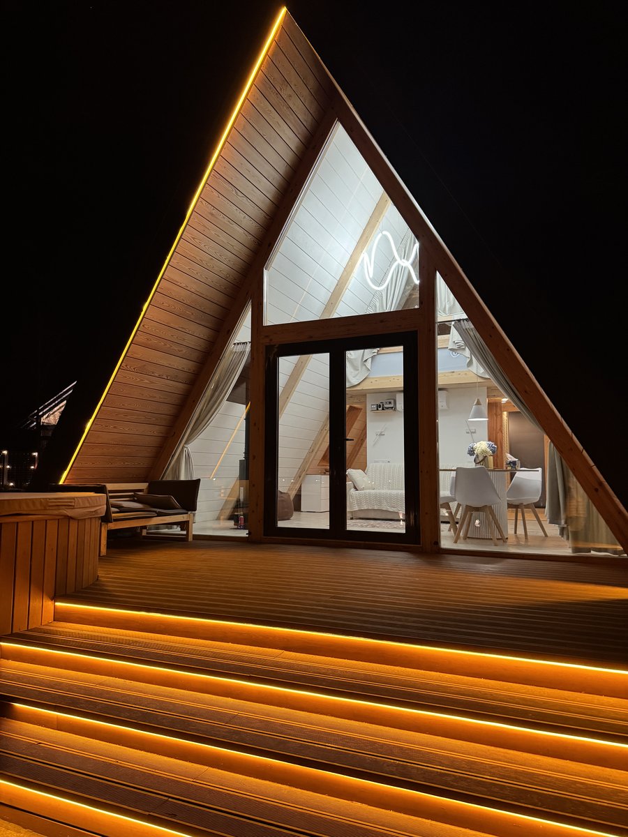 A-frame дом ночью со стороны террасы: тёплый свет внутри, LED-подсветка лестницы, луна в облаках